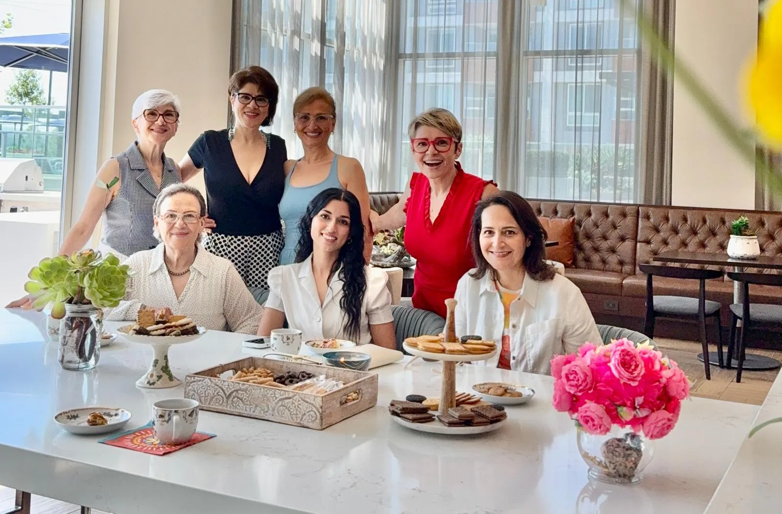 Seven women smiling together around a table set with tea, cookies, succulents, and pink roses